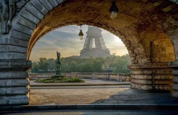 Eiffel Tower viewed through a stone archway of a bridge, with a statue and trees in the midground, under a hazy sky.