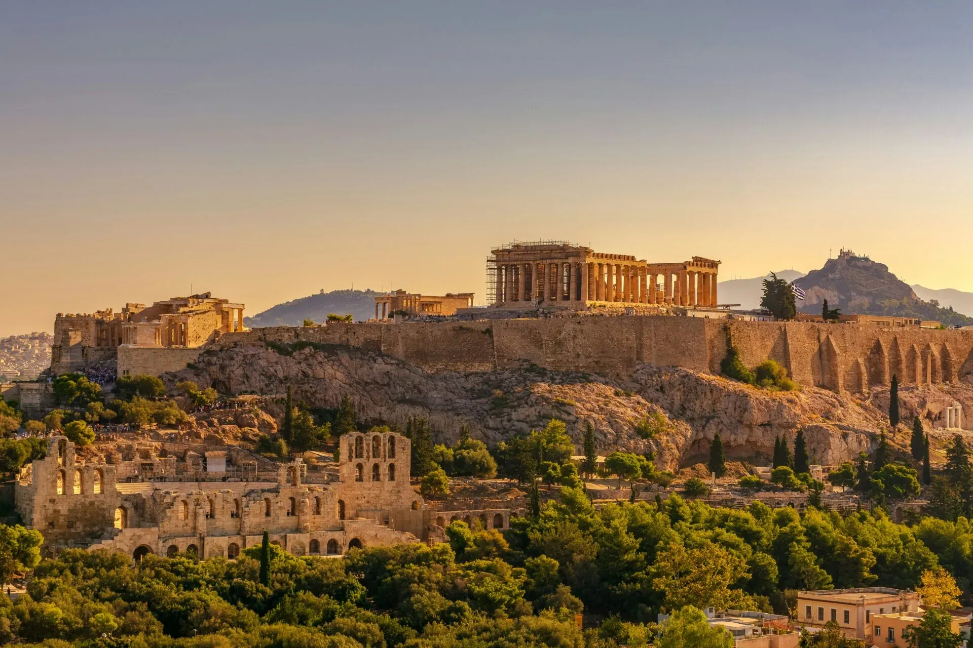 The Acropolis of Athens at sunset, showing the Parthenon, Erechtheion, and Odeon of Herodes Atticus, surrounded by trees.