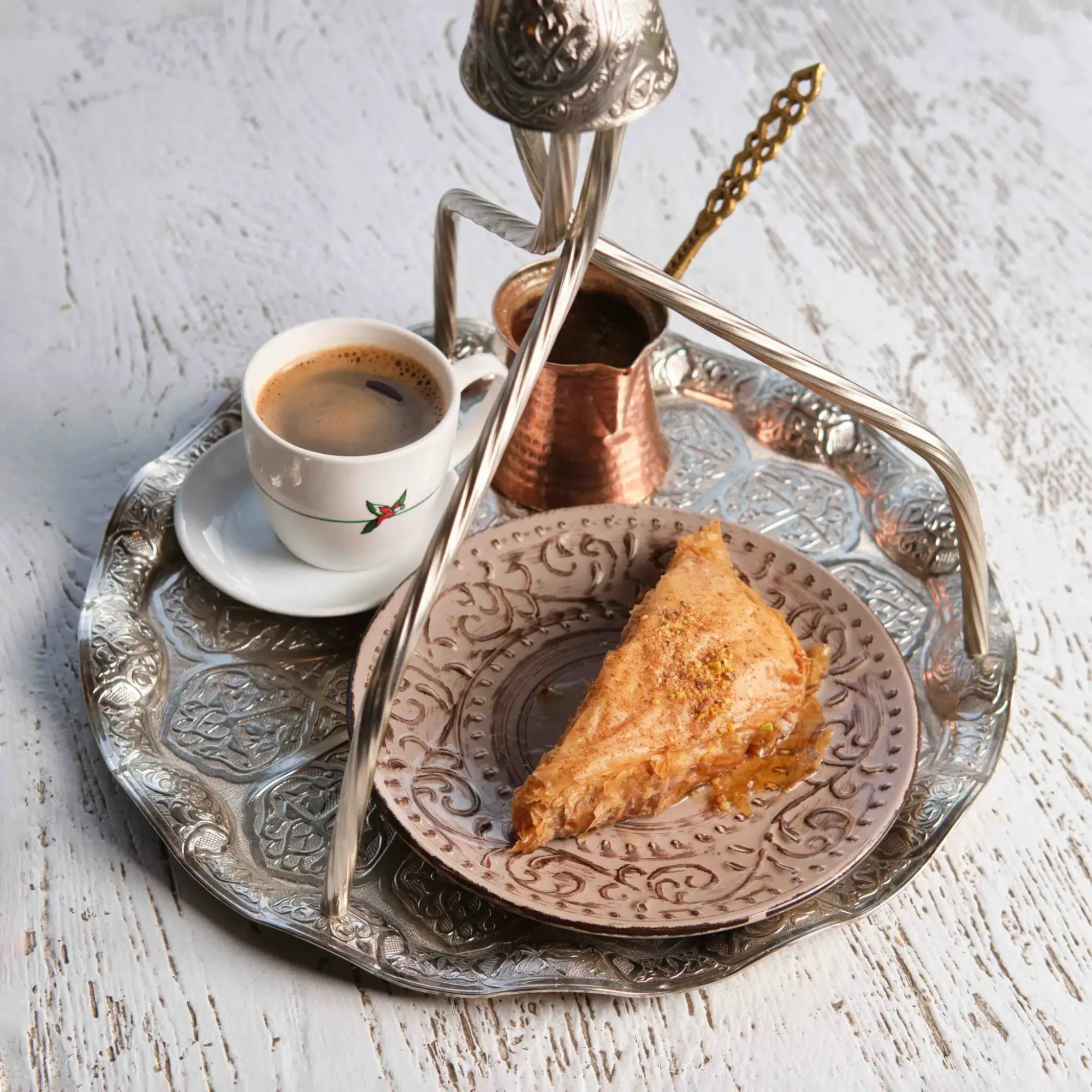 Hanging metal tray with a cup of Greek coffee, a copper pot, and a piece of baklava on a rustic white table.