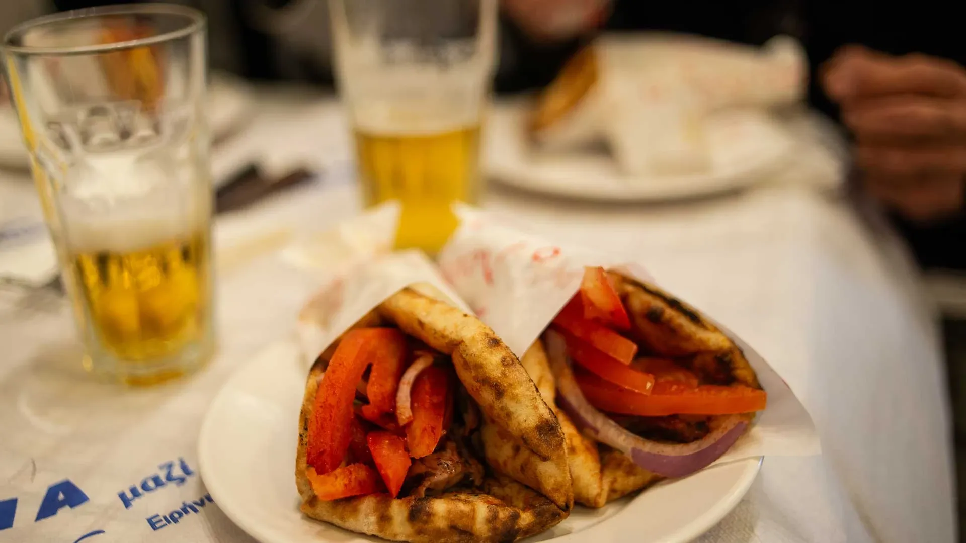 Two souvlaki in pita bread on a white plate, served with tomatoes and onions, with a glass of beer.