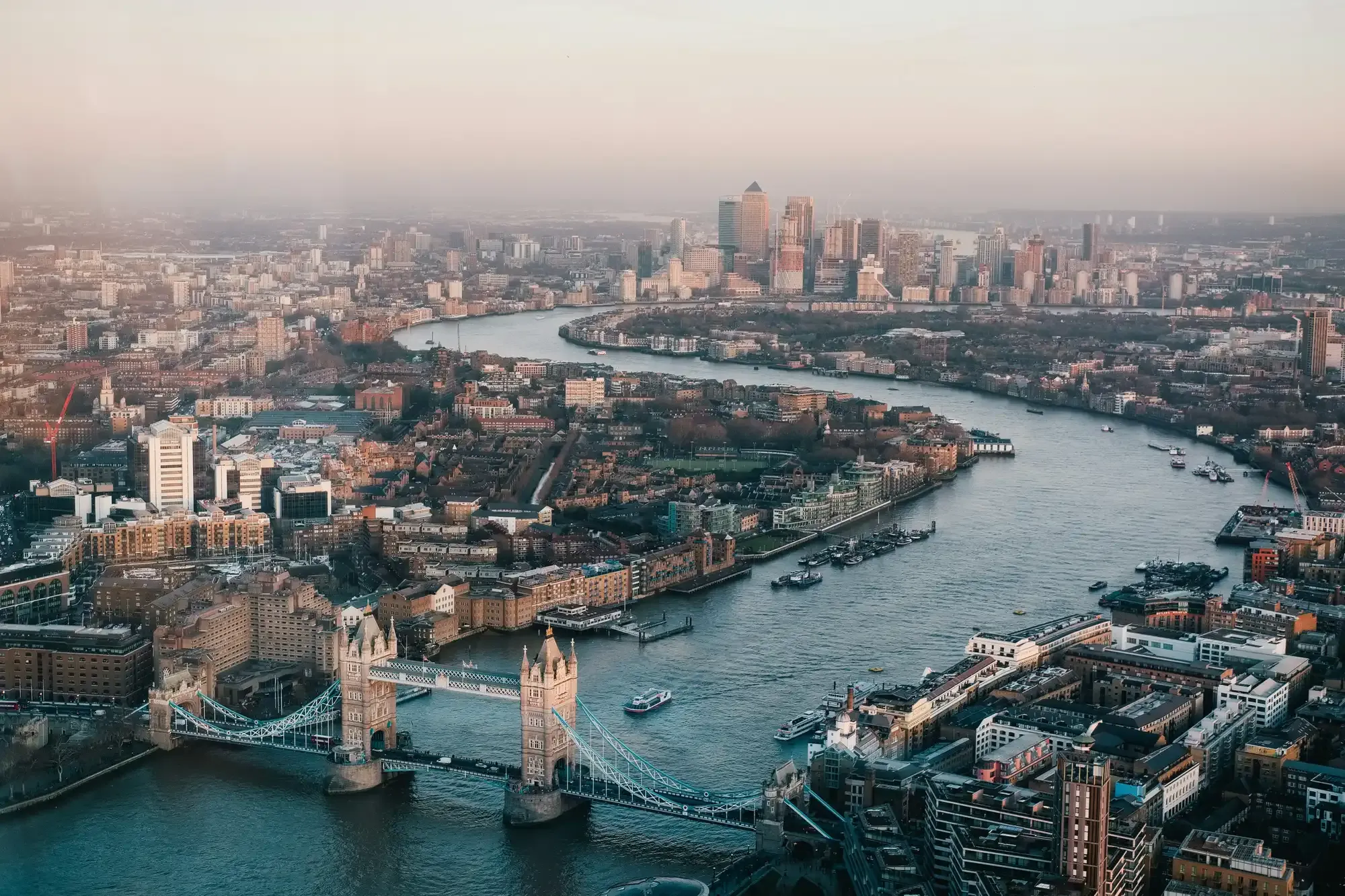 Aerial view of London with Tower Bridge and the River Thames, with the Canary Wharf skyline in the distance at dusk.