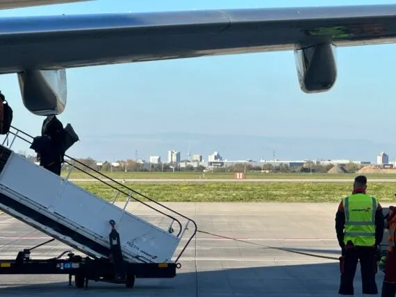 Underside of an airplane wing with passengers boarding via a mobile stair truck, airport ground crew, and a distant city skyl