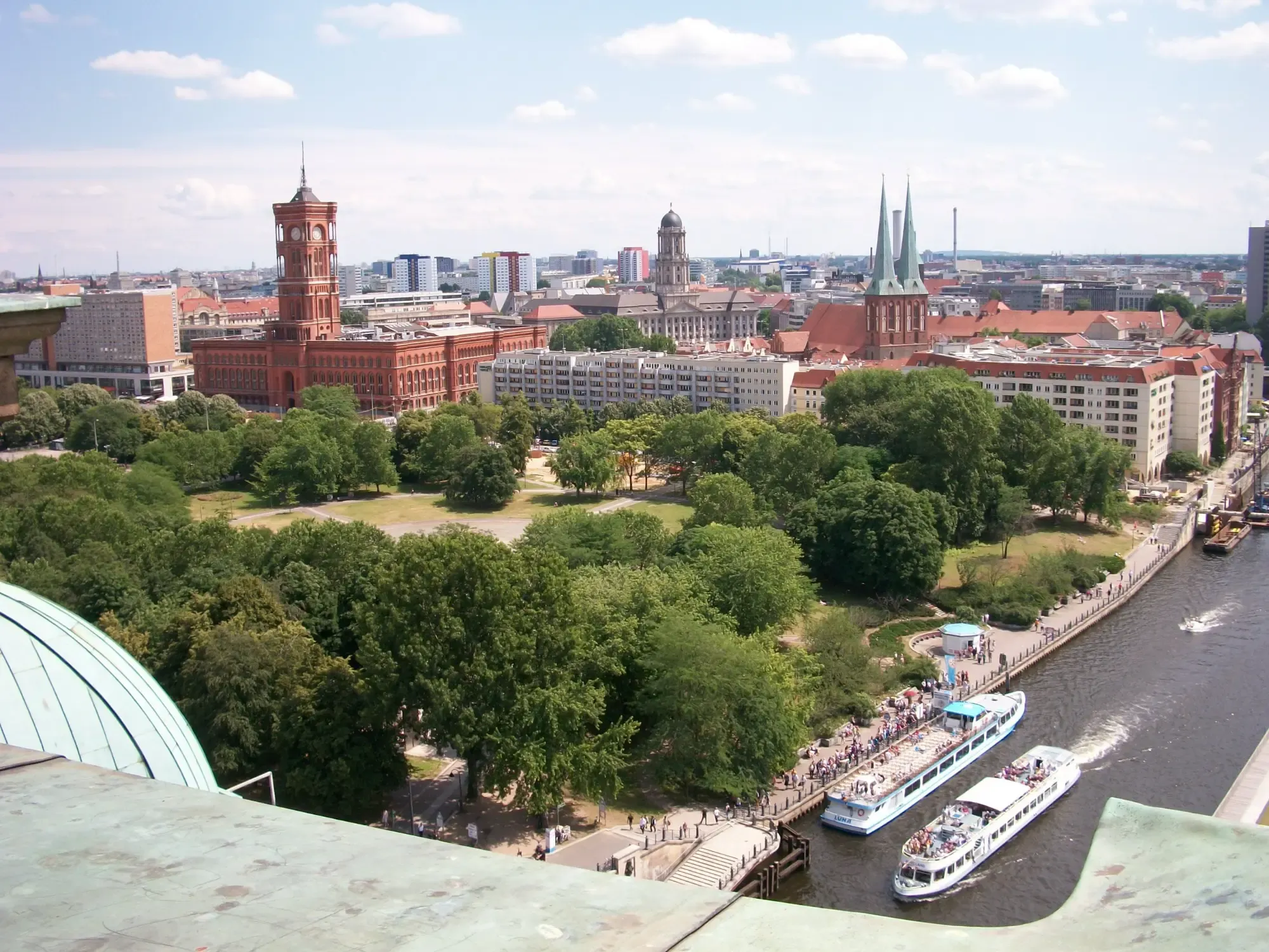 Aerial view of Berlin city center with the red brick Rotes Rathaus, green trees, the Spree River, and tour boats.