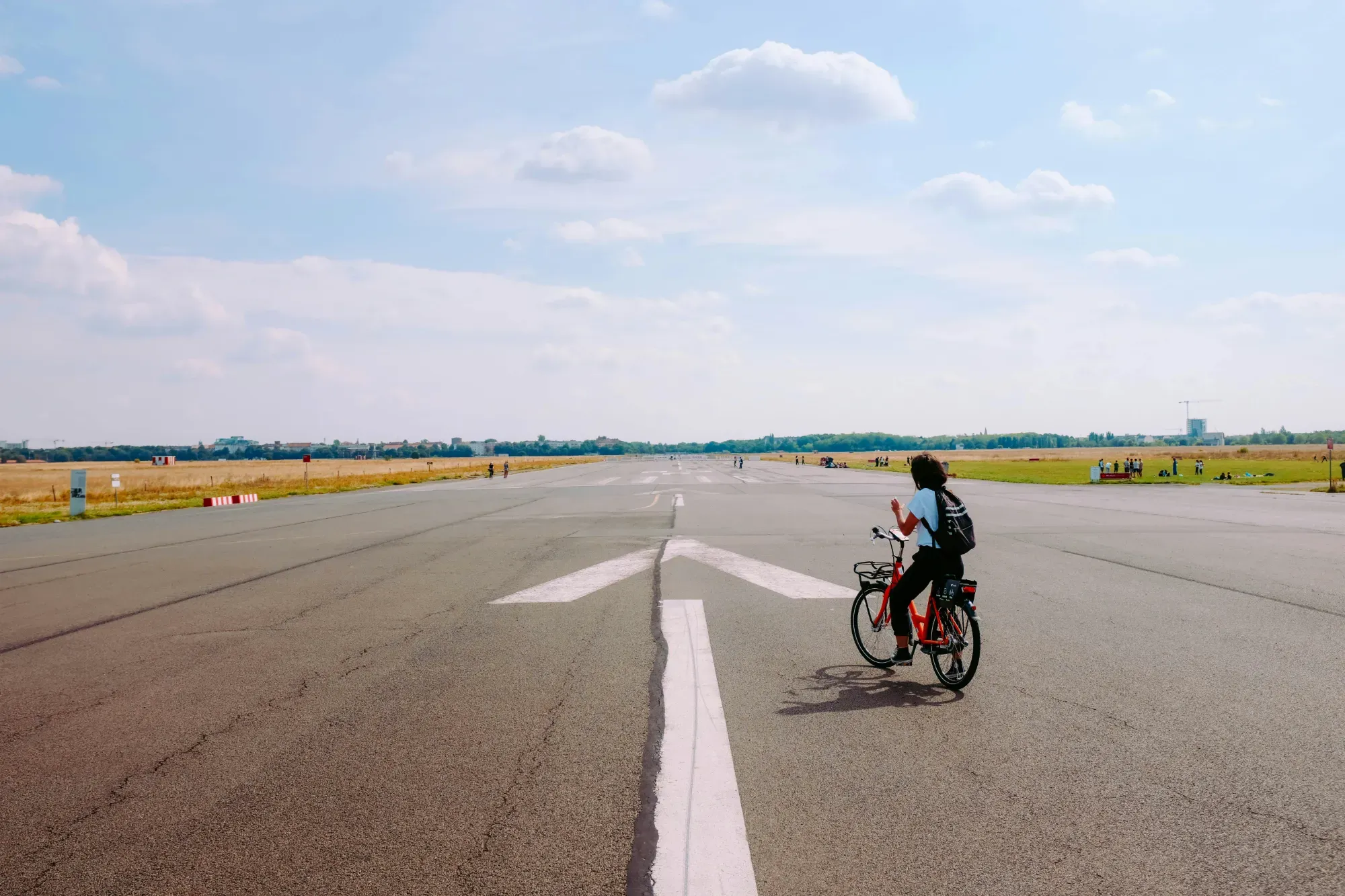 Person on a red bicycle on the paved runway of Tempelhofer Feld in Berlin, with fields and distant buildings under a blue sky