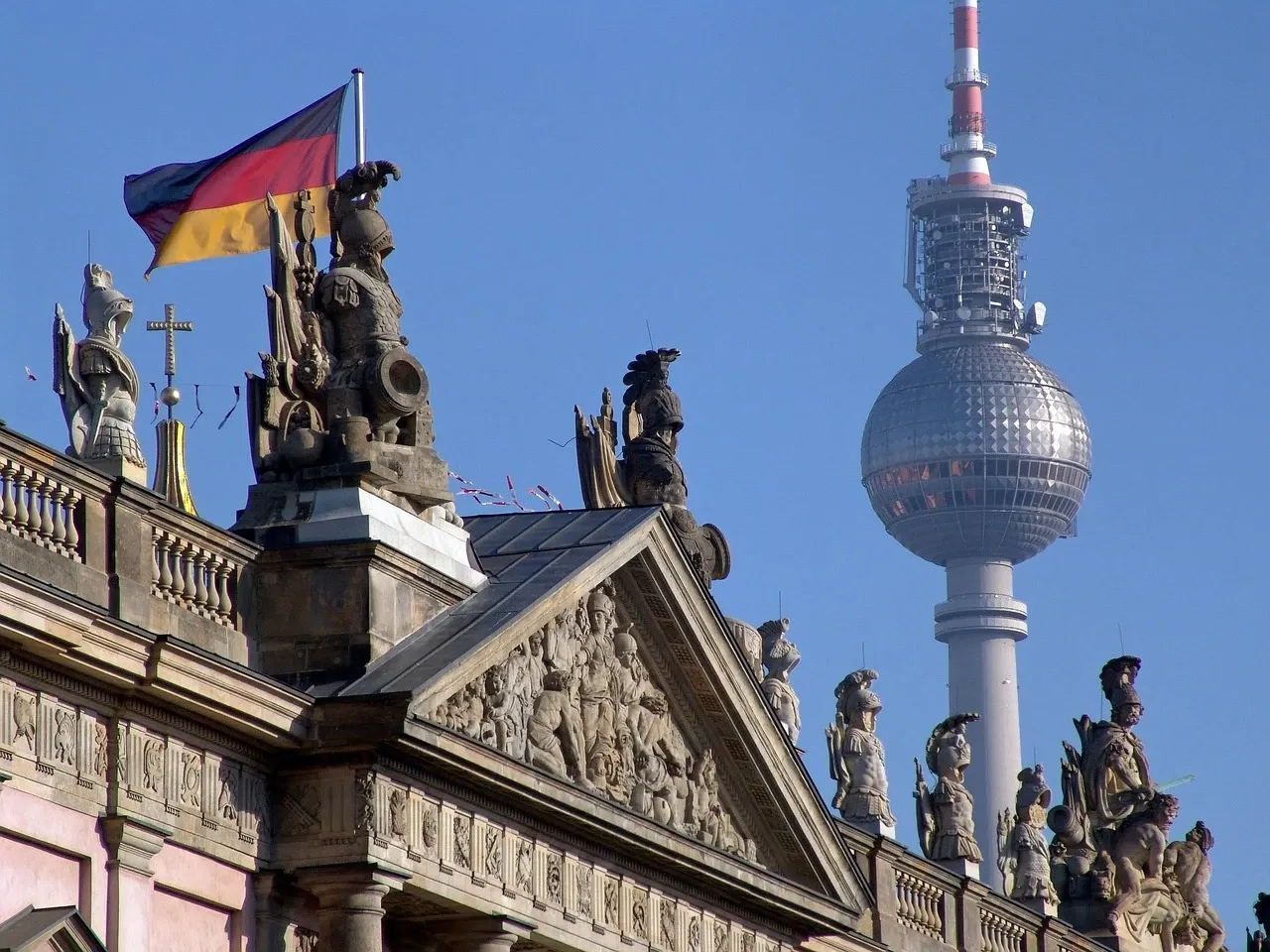 Zeughaus roof statues and German flag in the foreground with the iconic Berlin TV Tower rising behind against a blue sky.