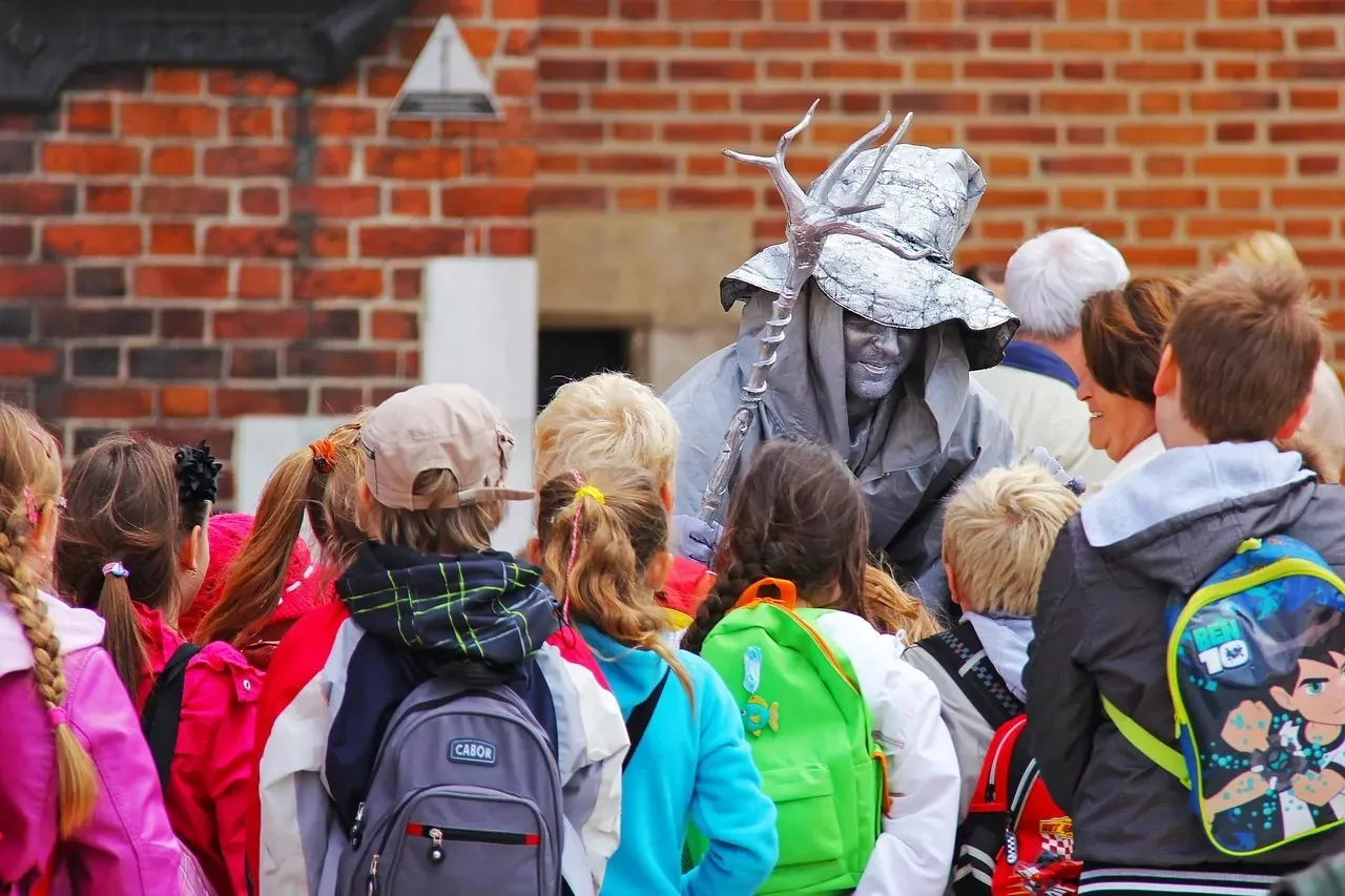 A silver-painted street performer in a hat and cloak interacts with a group of children and adults against a brick wall.