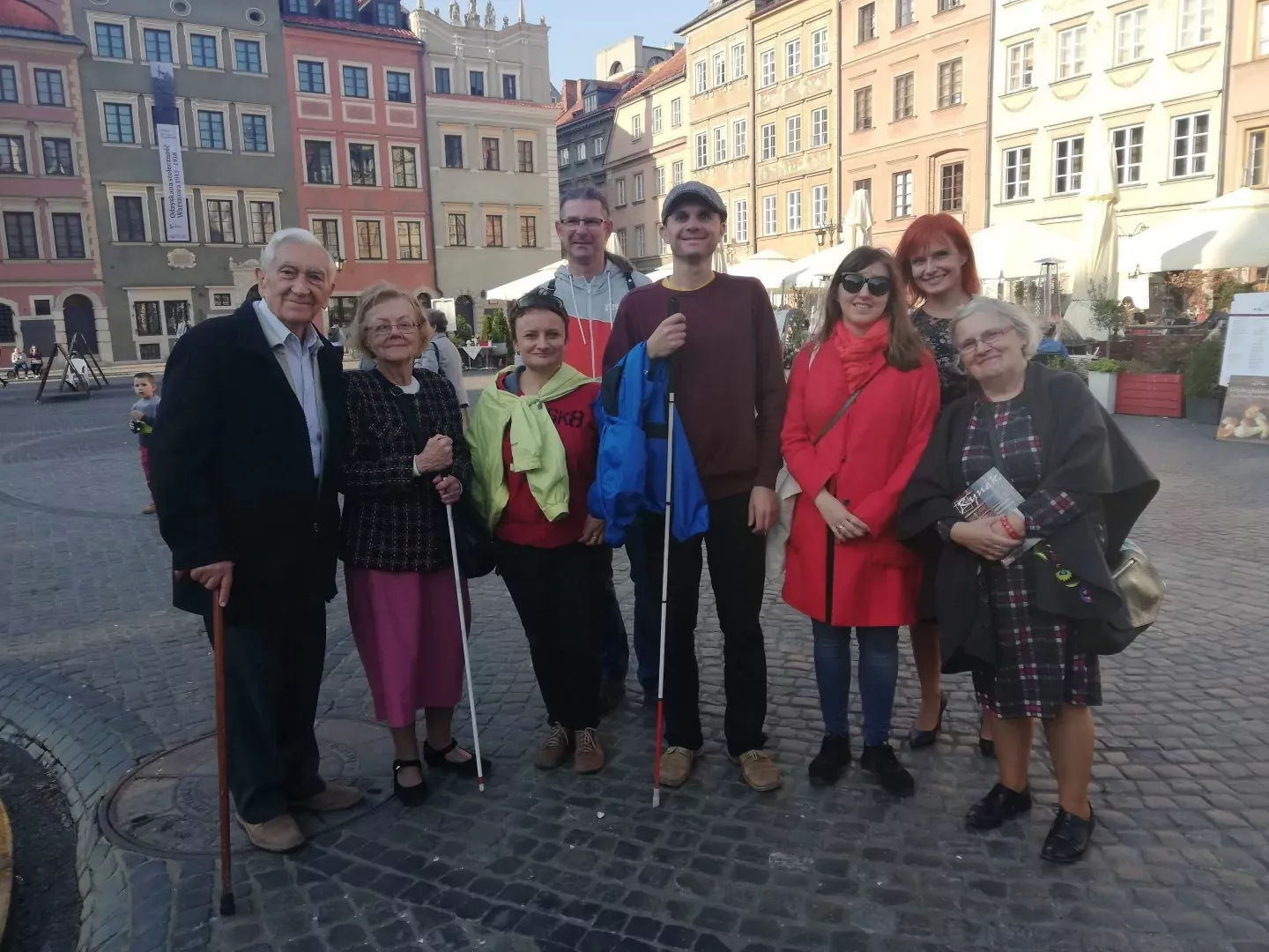 A diverse group of people, including some with white canes and a walking stick, stand on a cobblestone square surrounded by h
