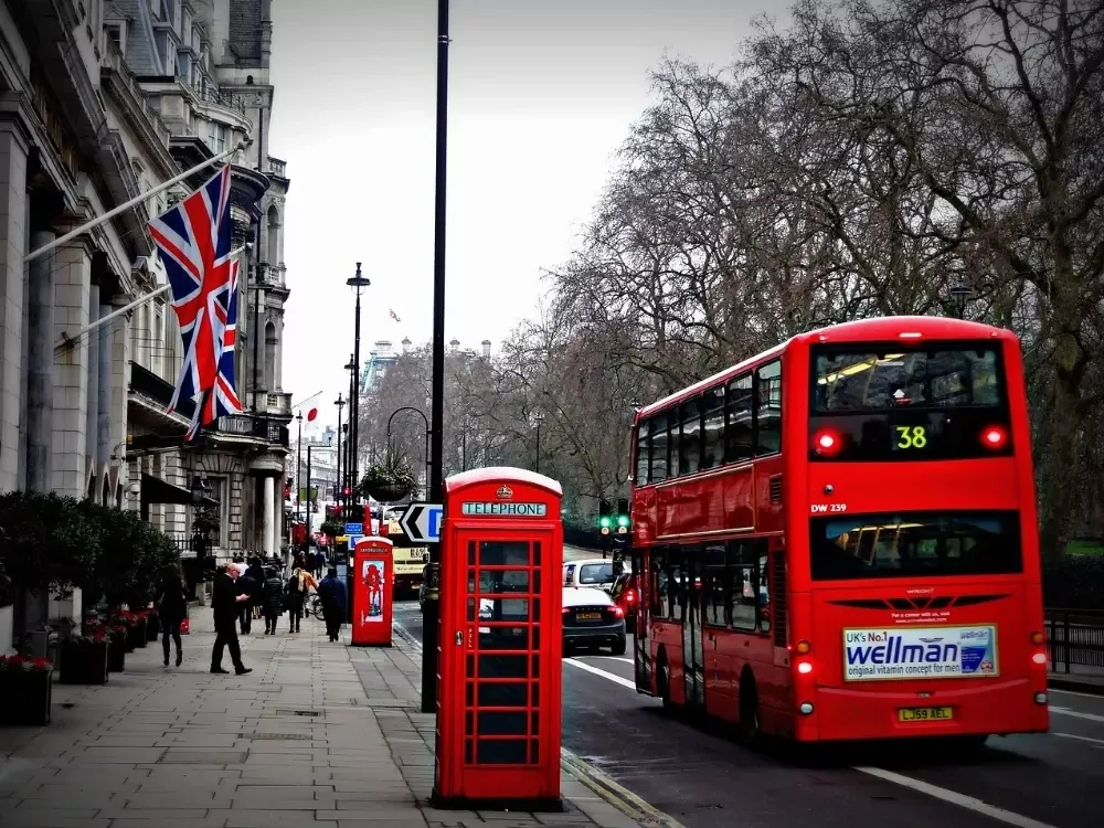 Red double-decker bus on a street next to a red phone booth and a building flying a Union Jack flag in London.