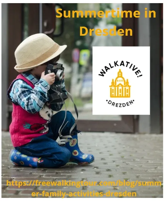 A small child in a hat and rain boots sits on a pavement, holding a vintage camera up to their eye.