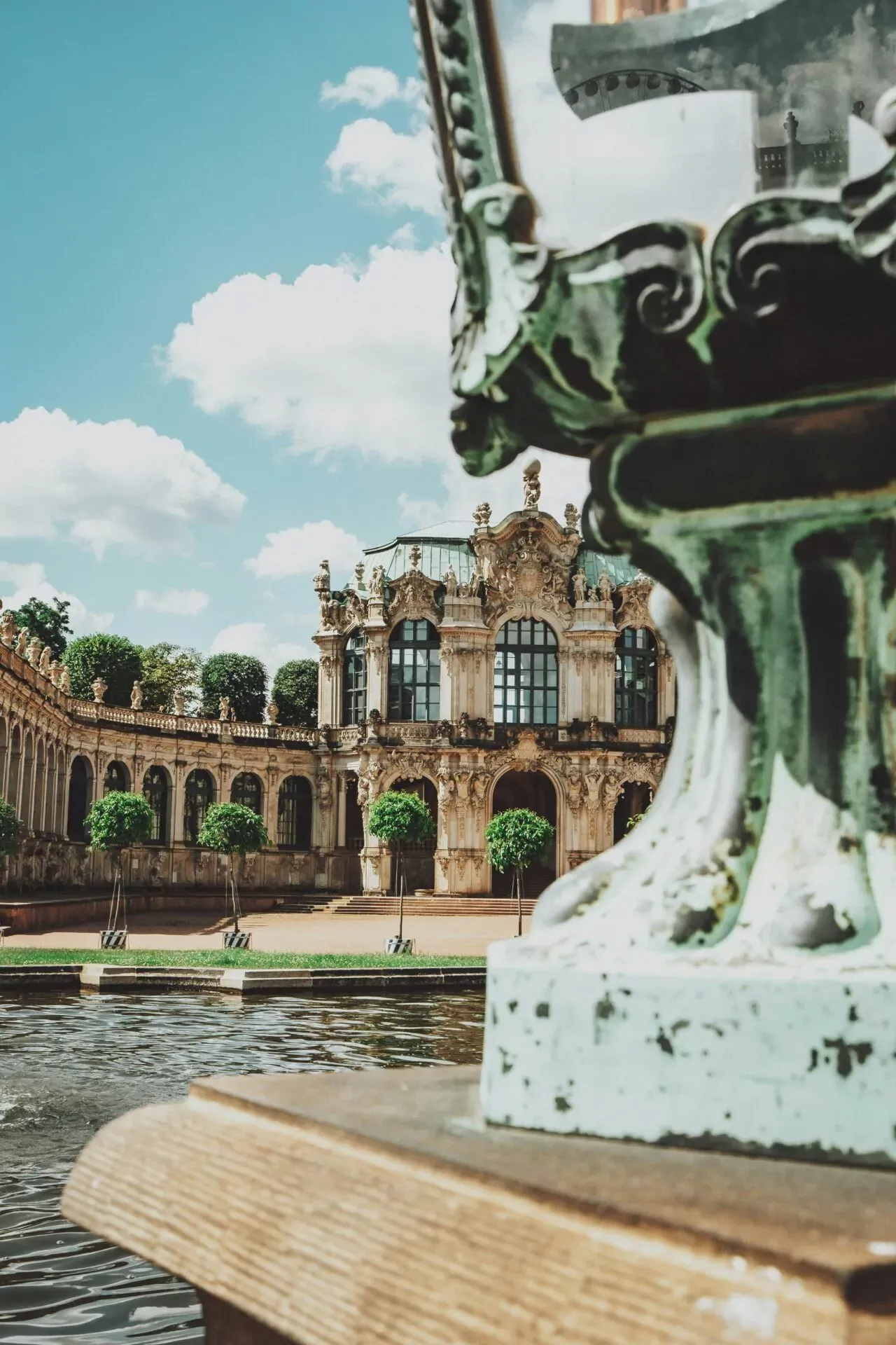 Foreground decorative metalwork by a pond, with the ornate Baroque architecture of the Zwinger Palace and arched galleries be