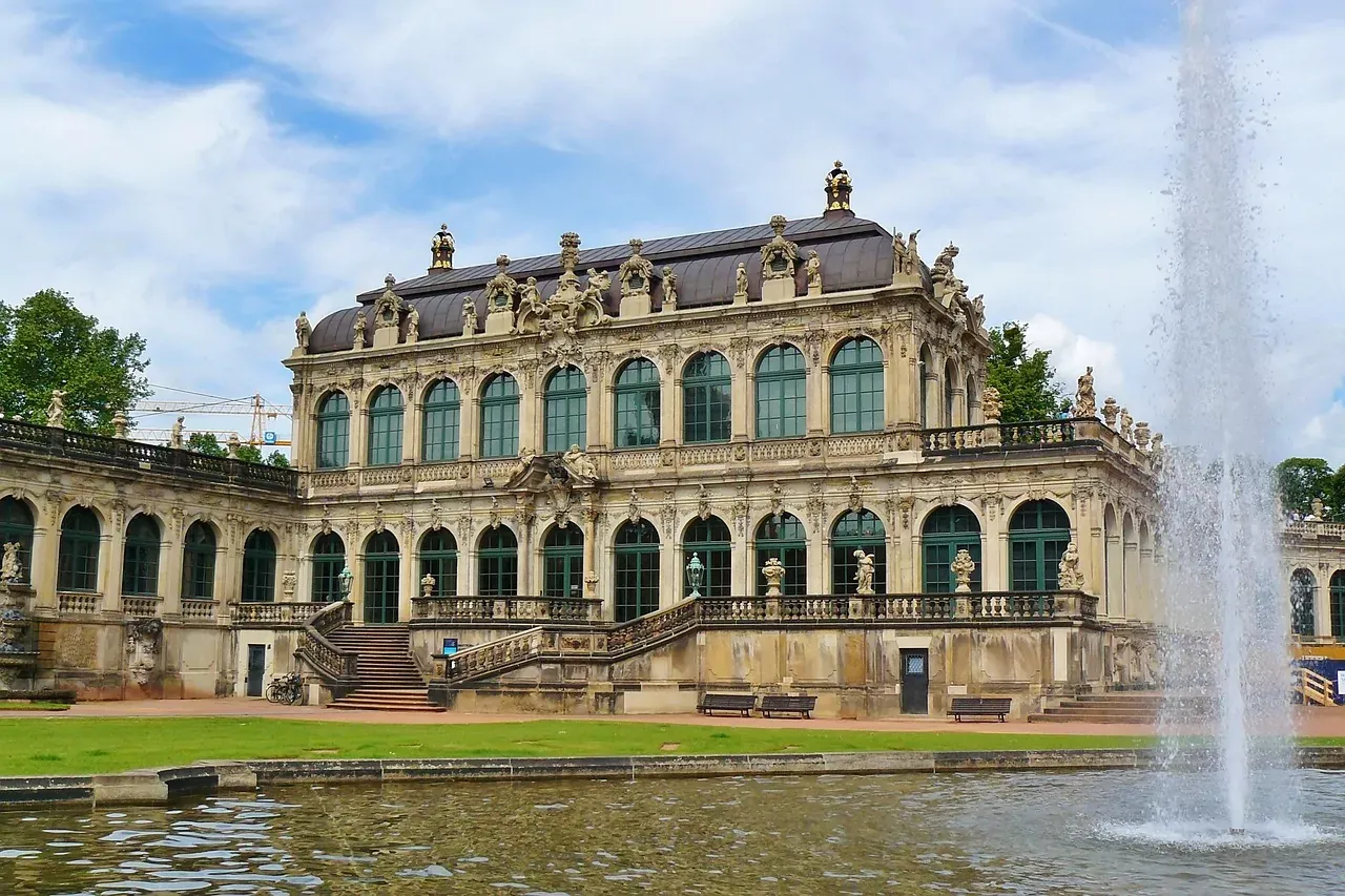 Ornate Baroque Zwinger Palace in Dresden with large arched windows, green roofs, and a fountain spraying water into a reflect