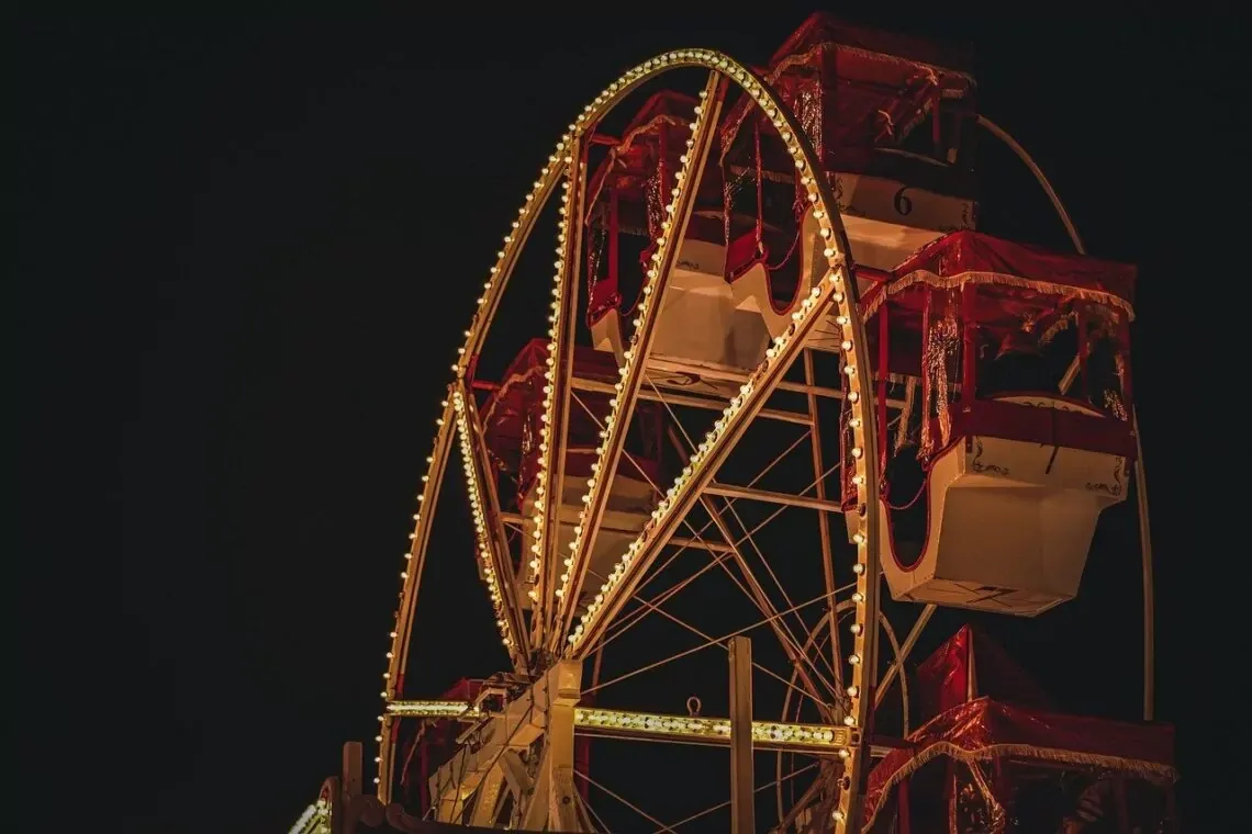Section of a Ferris wheel illuminated by warm lights at night against a dark sky. The passenger cabins are covered in red fab