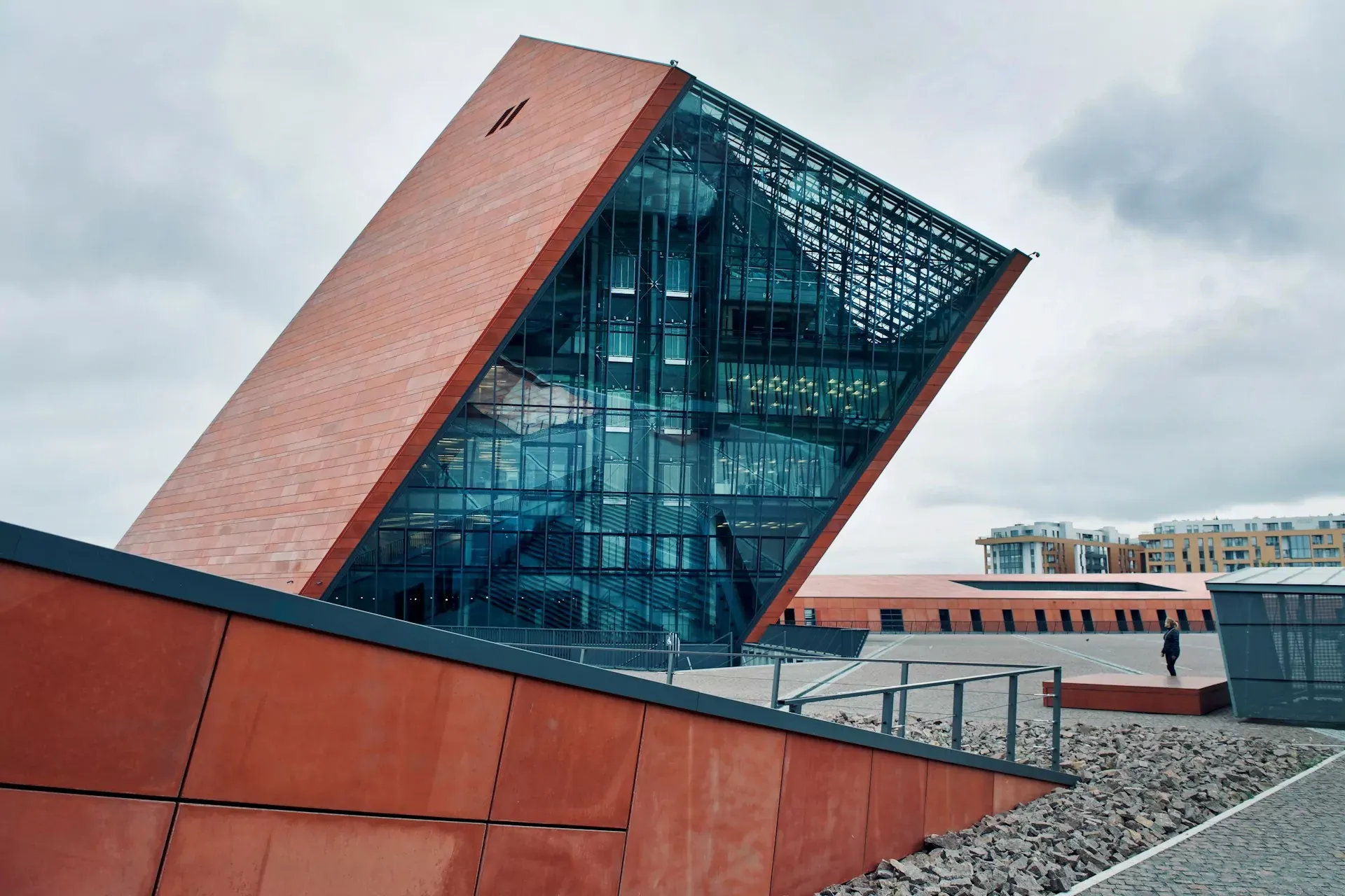 Angular, modern building with reddish-brown panels and large glass facade under an overcast sky in Gdansk. A person stands in