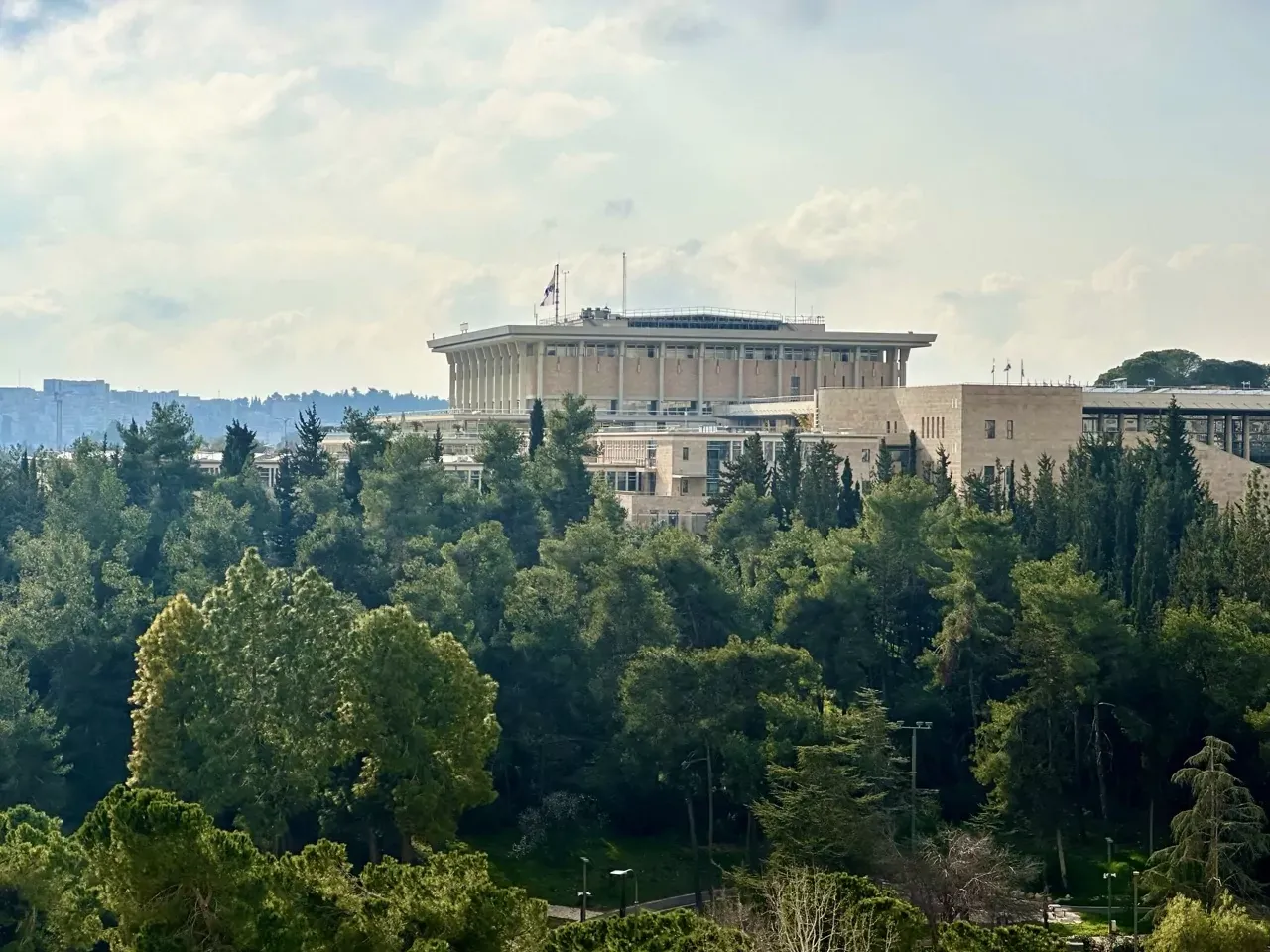 Large modern building with columns, partially hidden by dense green trees, under a cloudy sky.