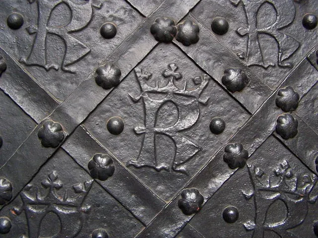Close-up of dark, ornate metal door with repeating crowned "R" monograms and decorative rivets in a diamond pattern.