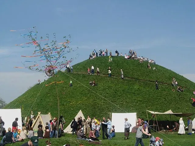 Crowd gathers on a grassy mound with a decorated pole and tents at its base under a blue sky in Krakow.