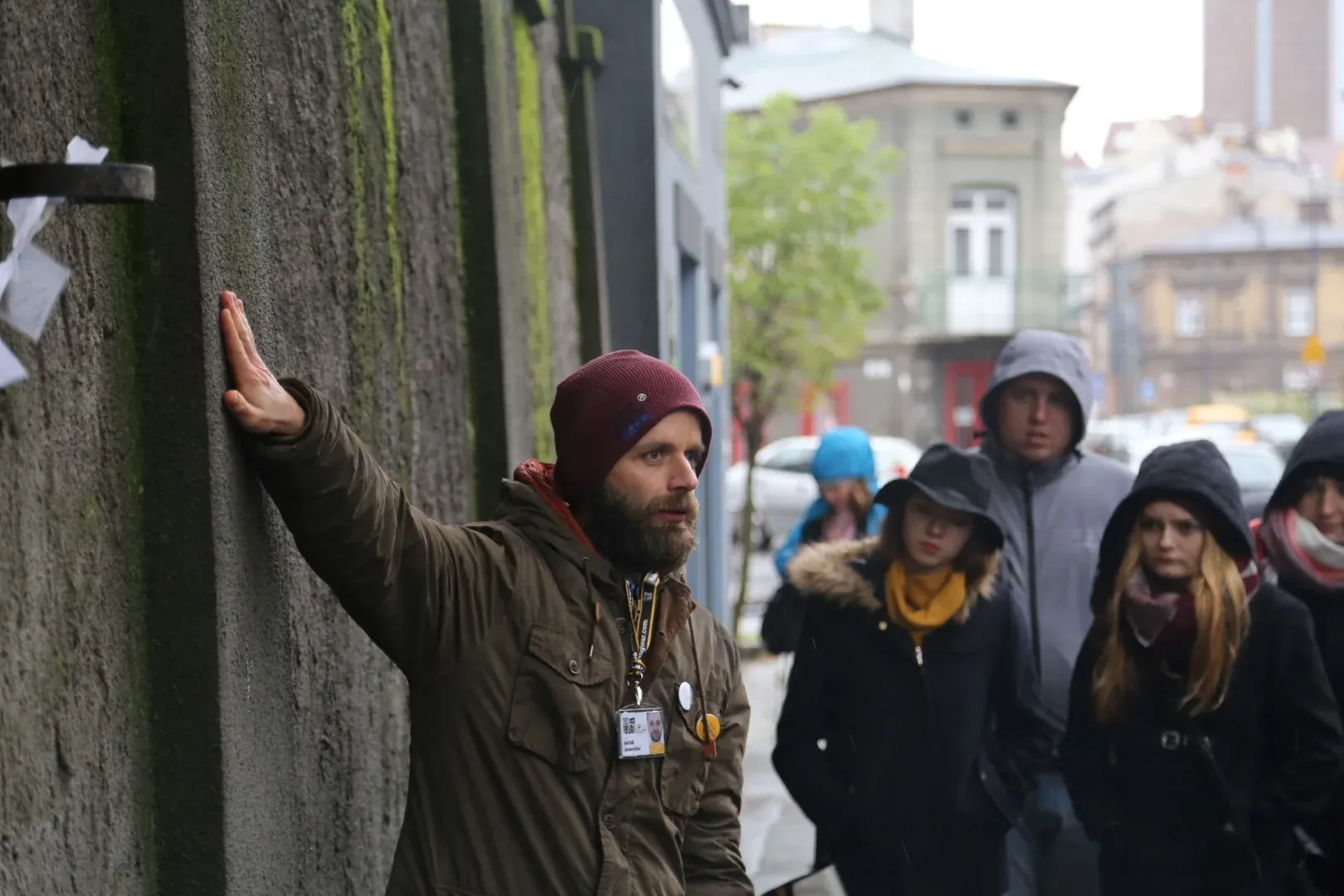 Bearded tour guide in a maroon beanie touches a mossy concrete wall while leading a group of people on a street.