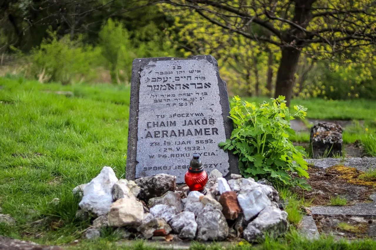 Weathered grey gravestone with Hebrew and Polish text, a red memorial candle, and small stones on grassy ground. Trees in bac