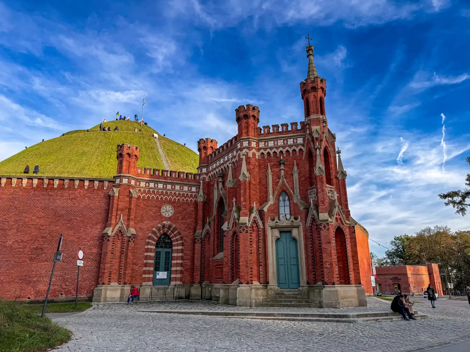 A red brick, Gothic Revival chapel in front of the grassy Kościuszko Mound in Krakow, under a blue sky.