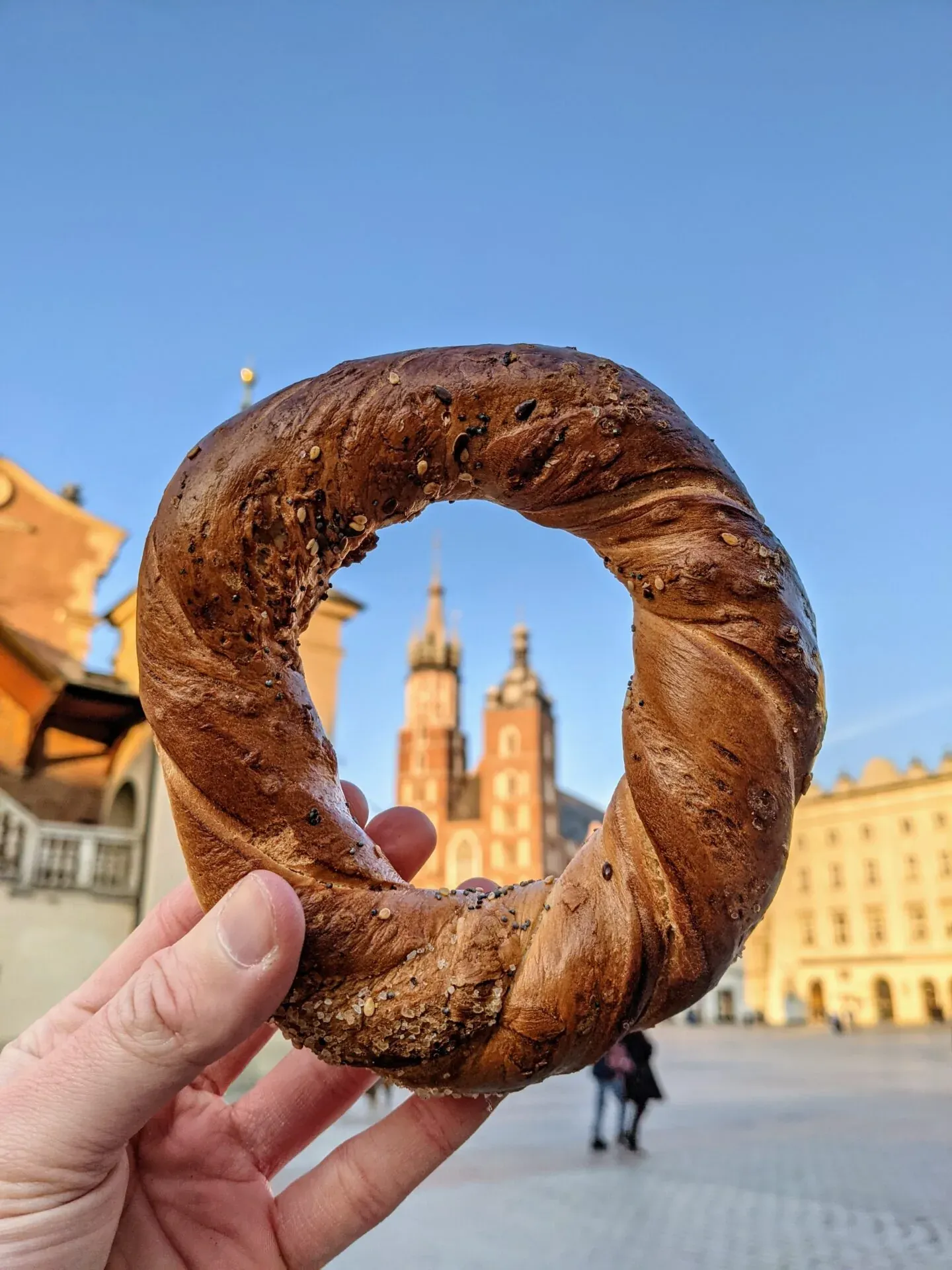 A hand holds a sesame and poppy seed obwarzanek, with St. Mary's Basilica and buildings of Krakow's Main Market Square blurre