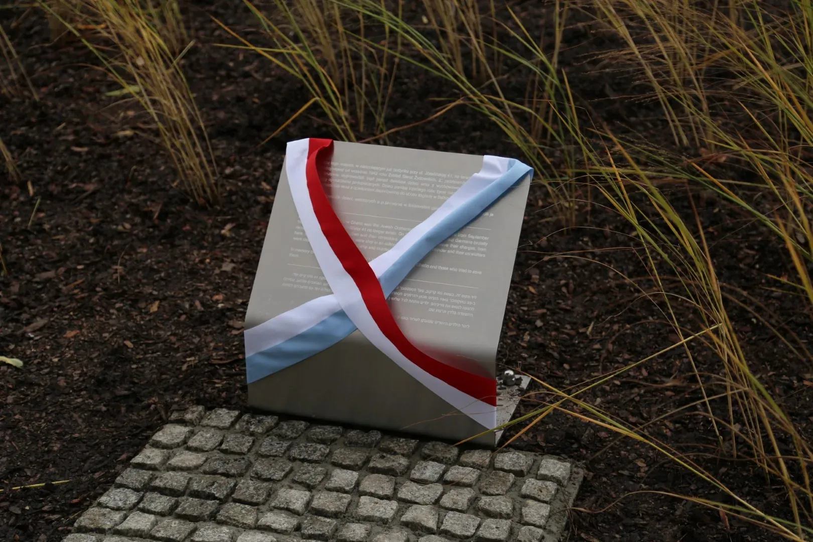 Grey memorial plaque with red, white, and light blue ribbons draped across it, resting on cobblestones near dry grass.