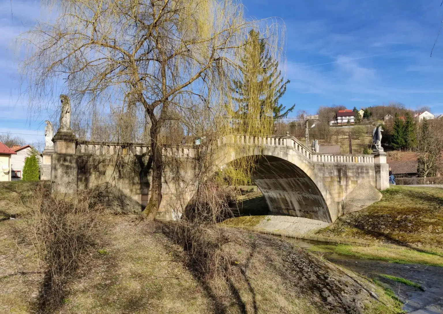 Stone arch bridge with a balustrade and statues, flanked by a bare willow tree and a small stream, under a blue sky.