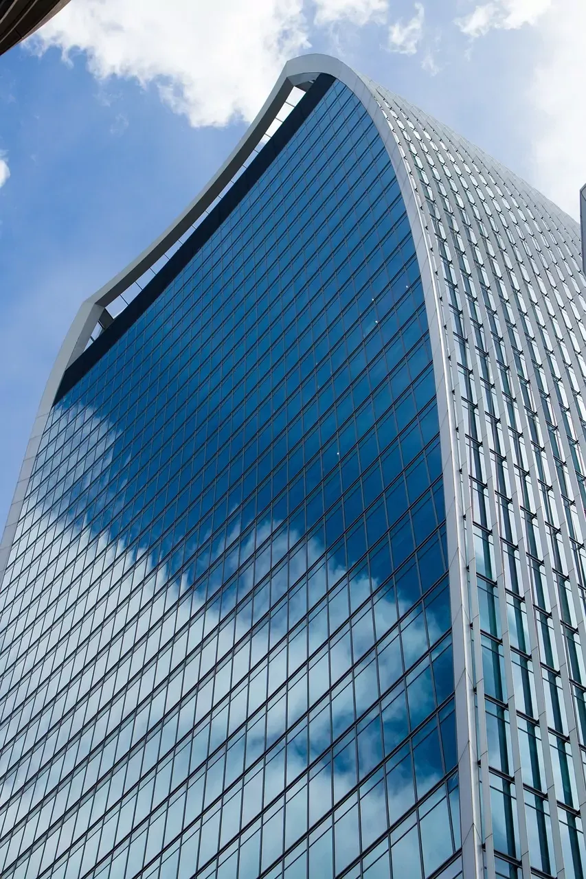 Curved glass facade of a modern skyscraper reflecting blue sky and white clouds, seen from a low angle.