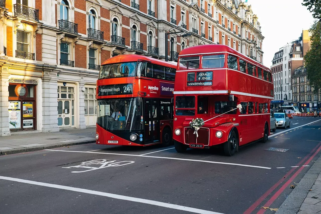 Two red double-decker buses on a London street. One is a public transport bus, the other a vintage bus decorated for a weddin