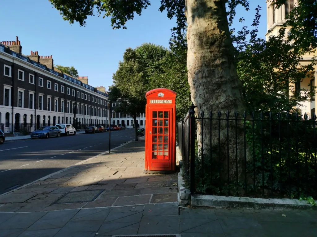 A bright red telephone box stands on a paved sidewalk next to a street with terraced houses and parked cars in London.