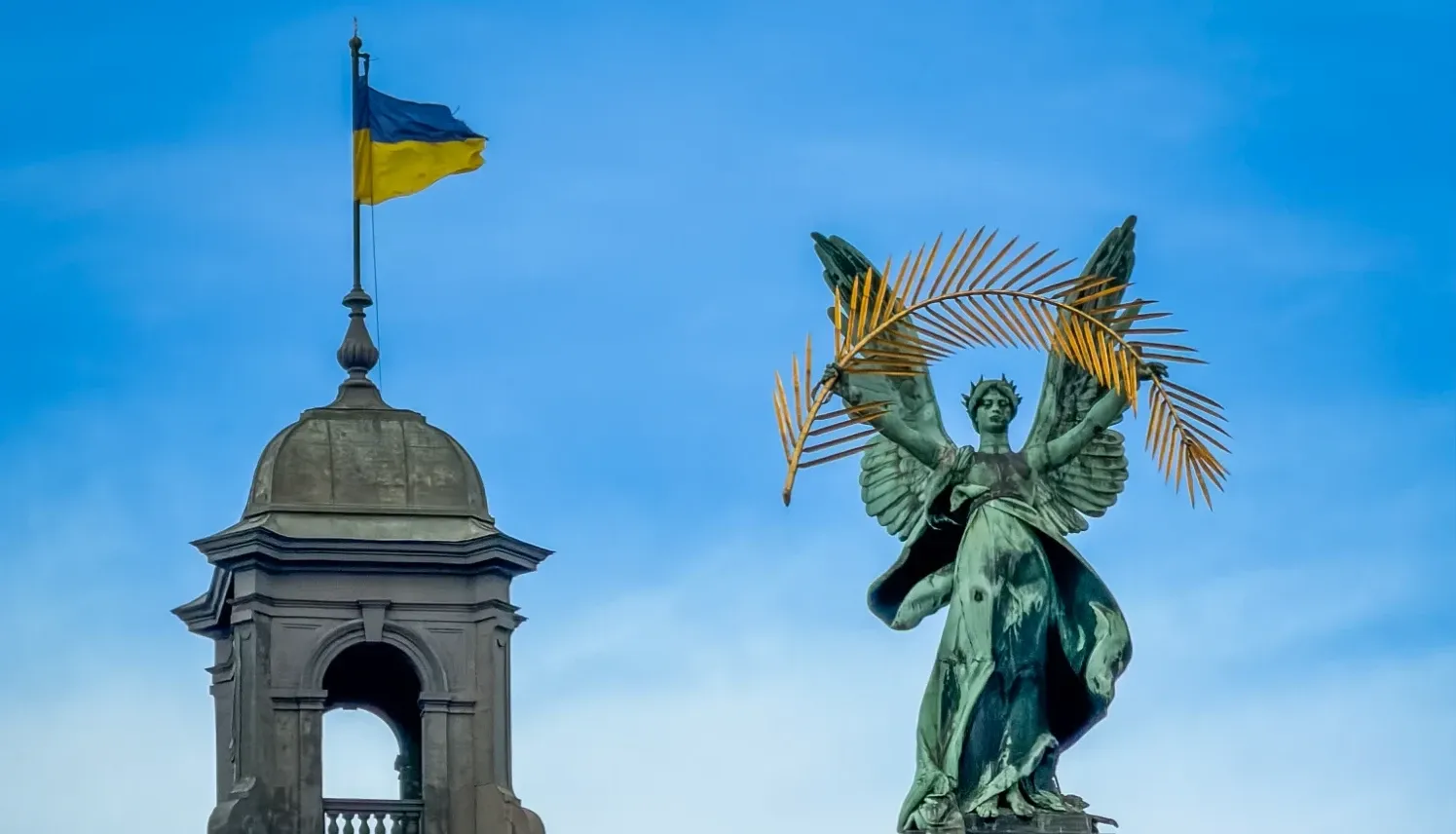 Bronze statue of Glory with a palm frond, next to a turret flying the Ukrainian flag against a blue sky.