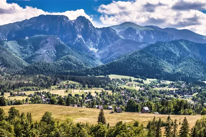 Vast mountain range with dense green forests covers the slopes, leading down to a valley with scattered houses and fields und