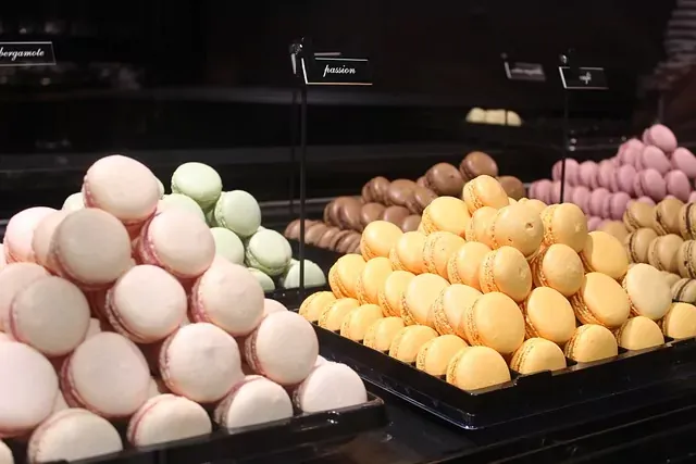 Piles of pink, green, yellow, brown, and purple macarons on black trays in a bakery display. Labels read 'bergamote' and 'pas