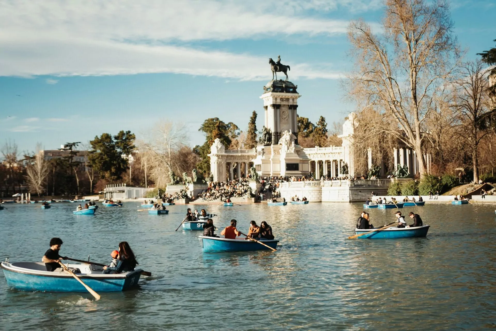 Many people in blue rowing boats on a lake with a grand stone monument and bare trees under a blue sky.