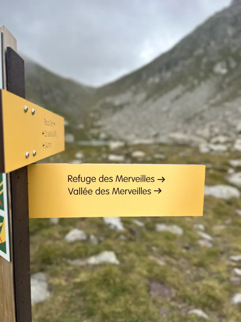 Yellow hiking sign pointing to Refuge des Merveilles and Vallée des Merveilles against a rocky mountain background.