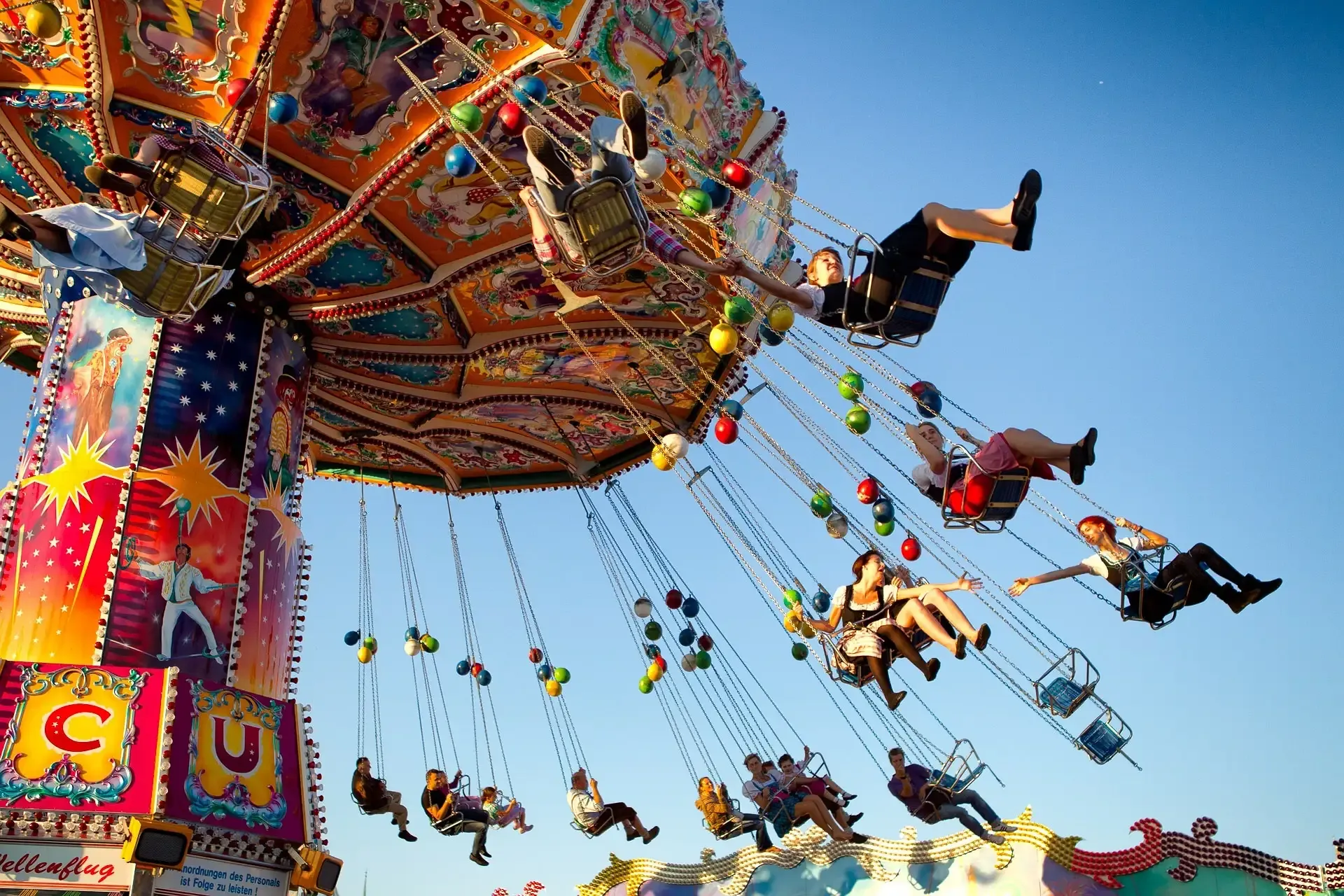 Colorful wave swinger carousel ride with multiple people suspended mid-air against a clear blue sky. The ride's decorated bas