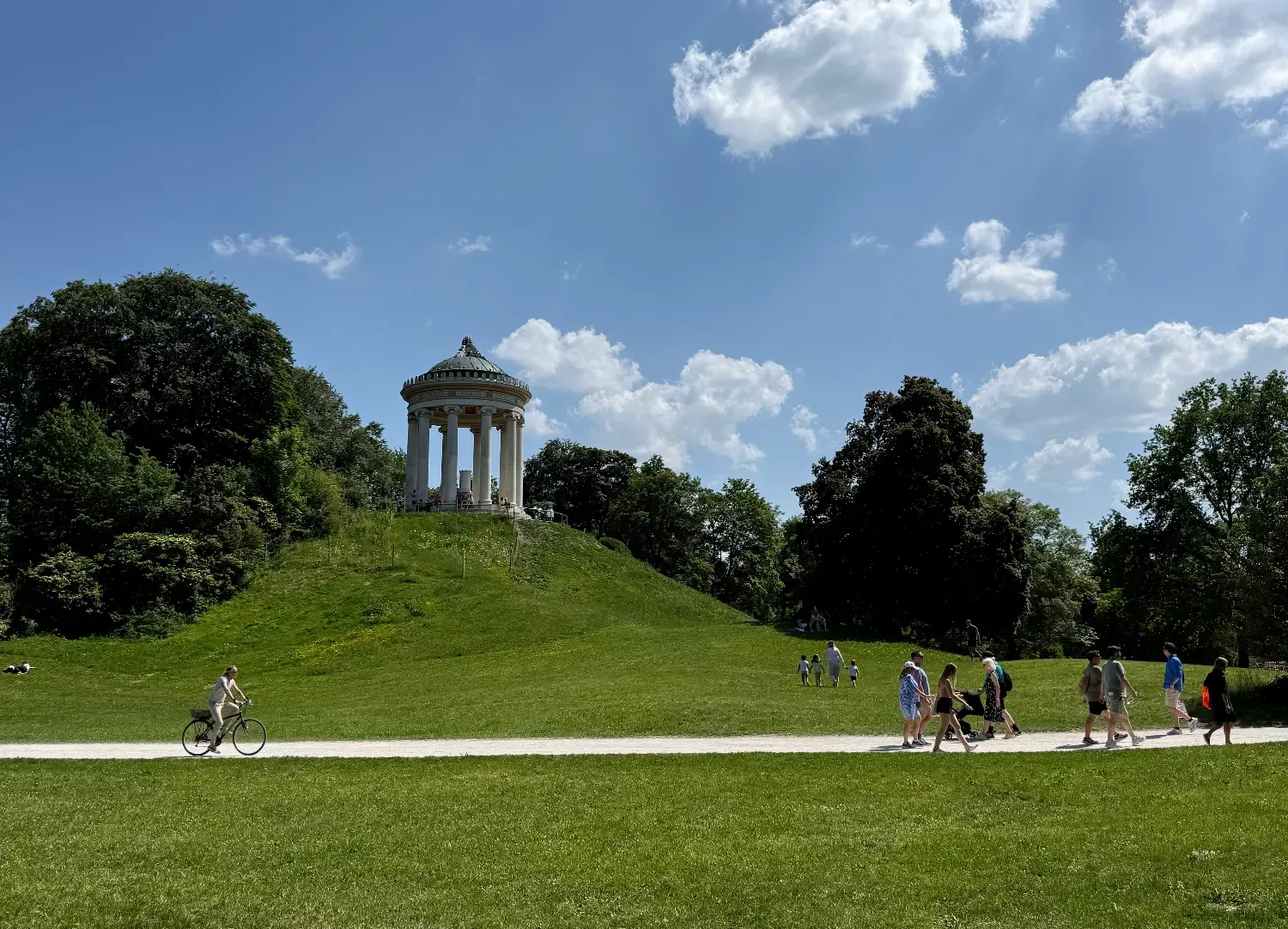 The Monopteros temple on a green hill in the English Garden, with people walking and cycling on the path below.