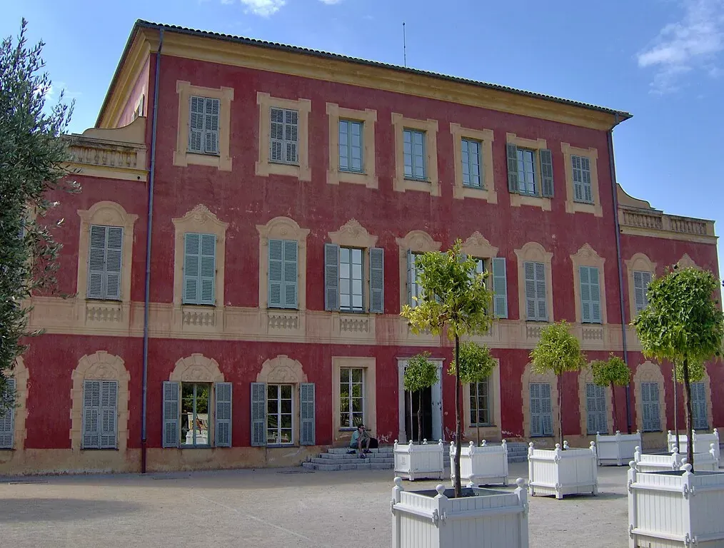Reddish-pink building with cream architectural details and light blue shutters. Several small trees in white planters are in