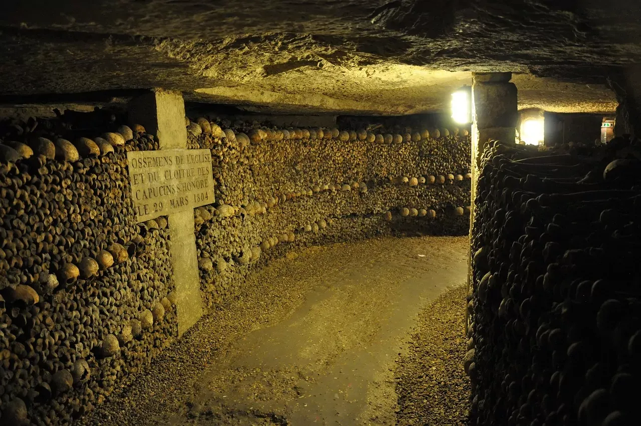 Underground tunnel with walls of human skulls and bones, illuminated by dim light. A stone sign is visible on the left.
