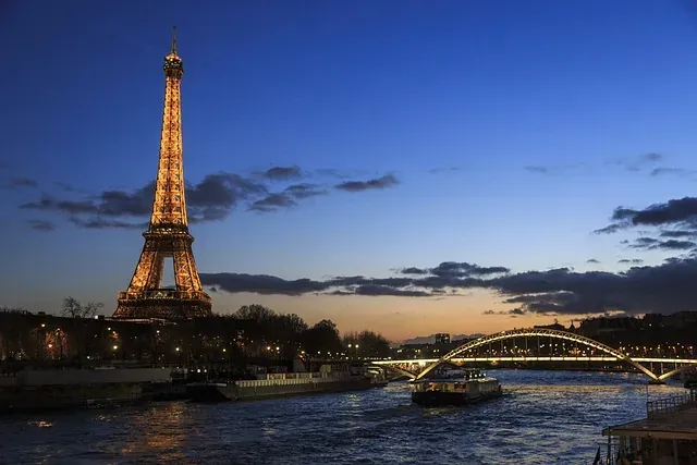 Illuminated Eiffel Tower and Pont de Bir-Hakeim bridge at dusk, with boats on the Seine River in Paris.