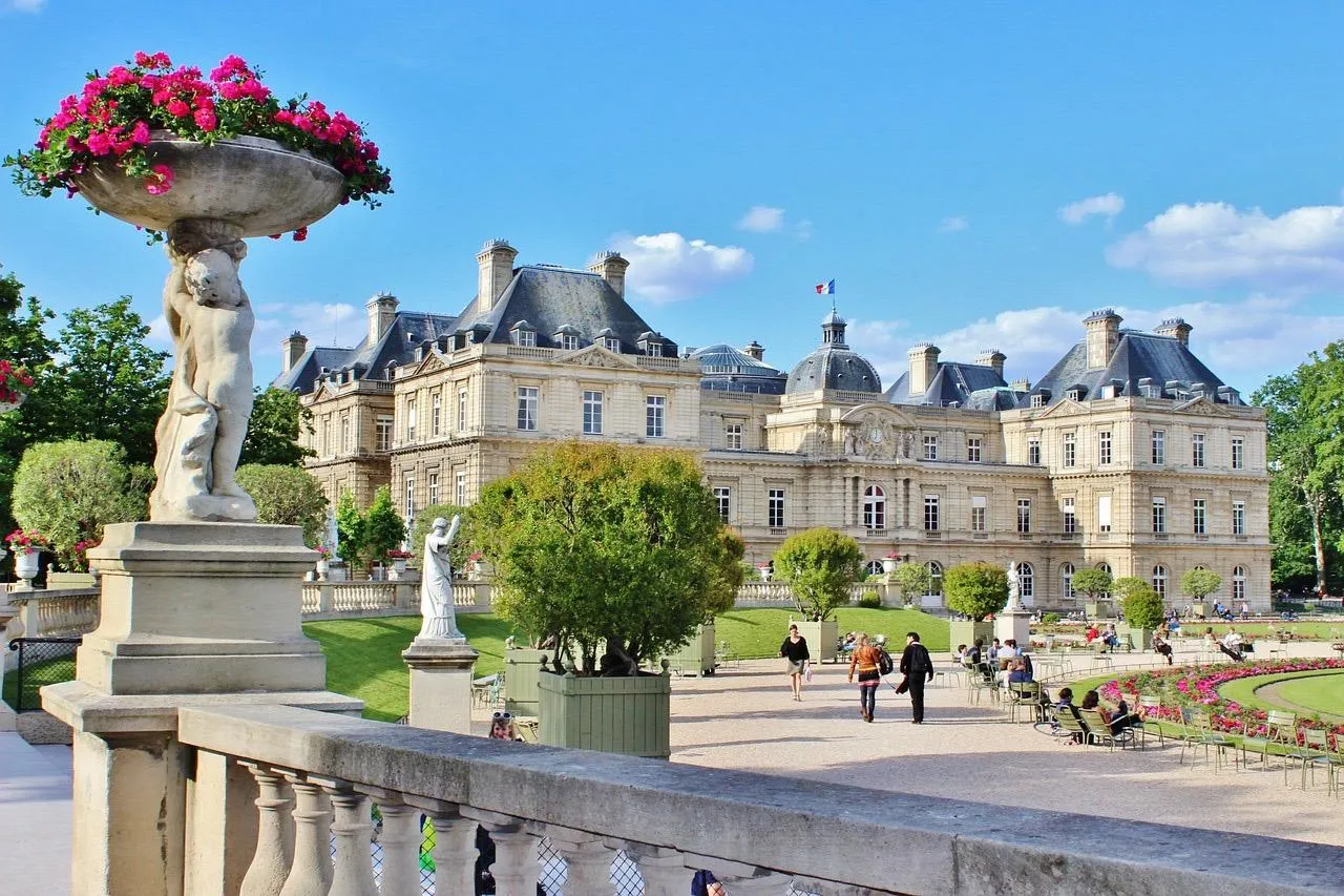 View of Luxembourg Palace from the gardens, with a large stone balustrade, statues, flowering plants, and people strolling.