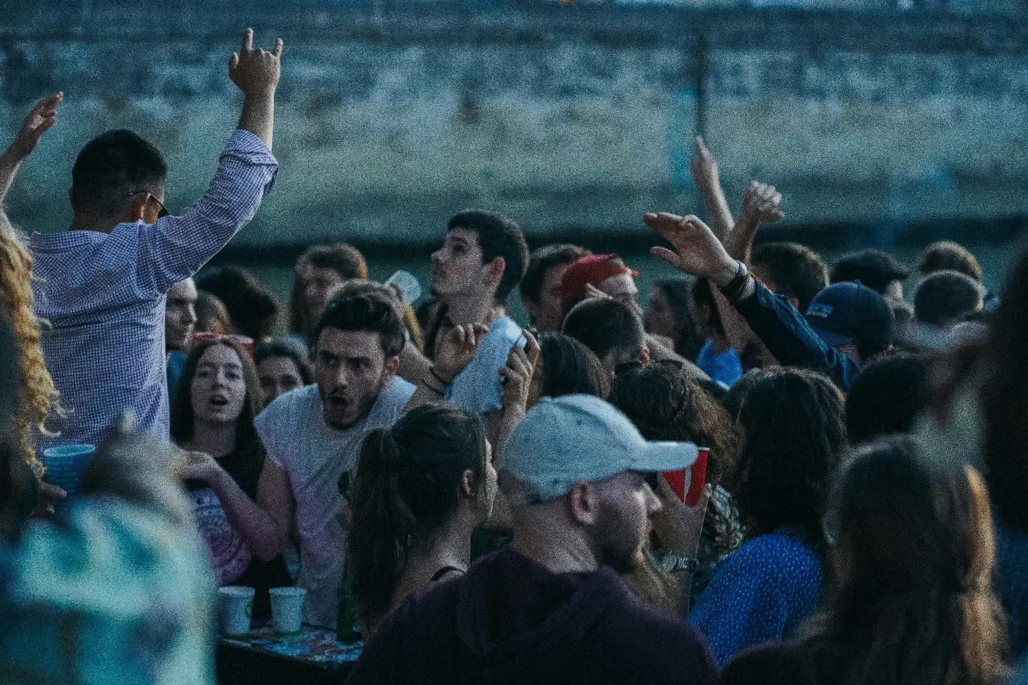 A dense crowd of young people at an outdoor evening event, with some raising their hands in the air.