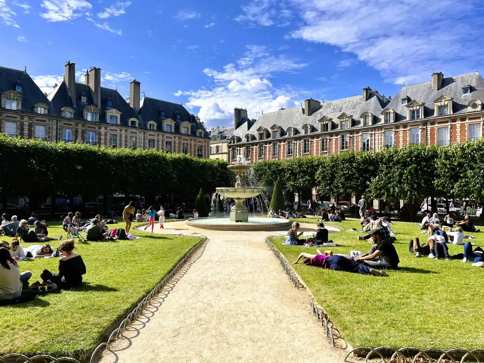 Place des Vosges in Paris with a central fountain, green lawns, people relaxing, and historic red brick buildings under a blu