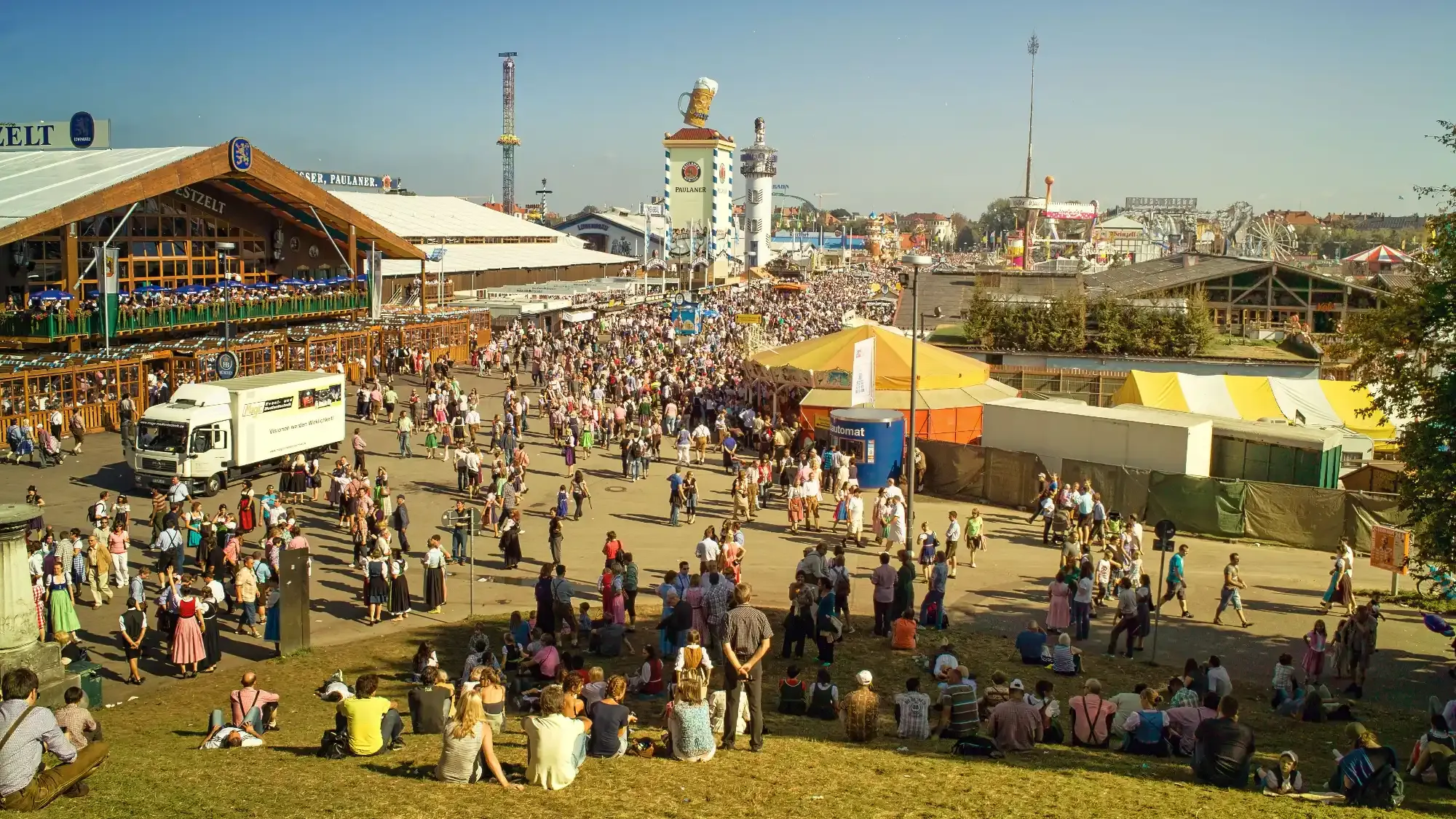 A sunny day at the Oktoberfest in Munich. Crowds of people mill between beer tents, rides, and stalls under a blue sky.