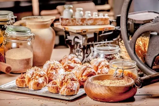 A silver tray holds several glazed croissants topped with nuts. Jars of grains and a vintage scale are in the blurred backgro