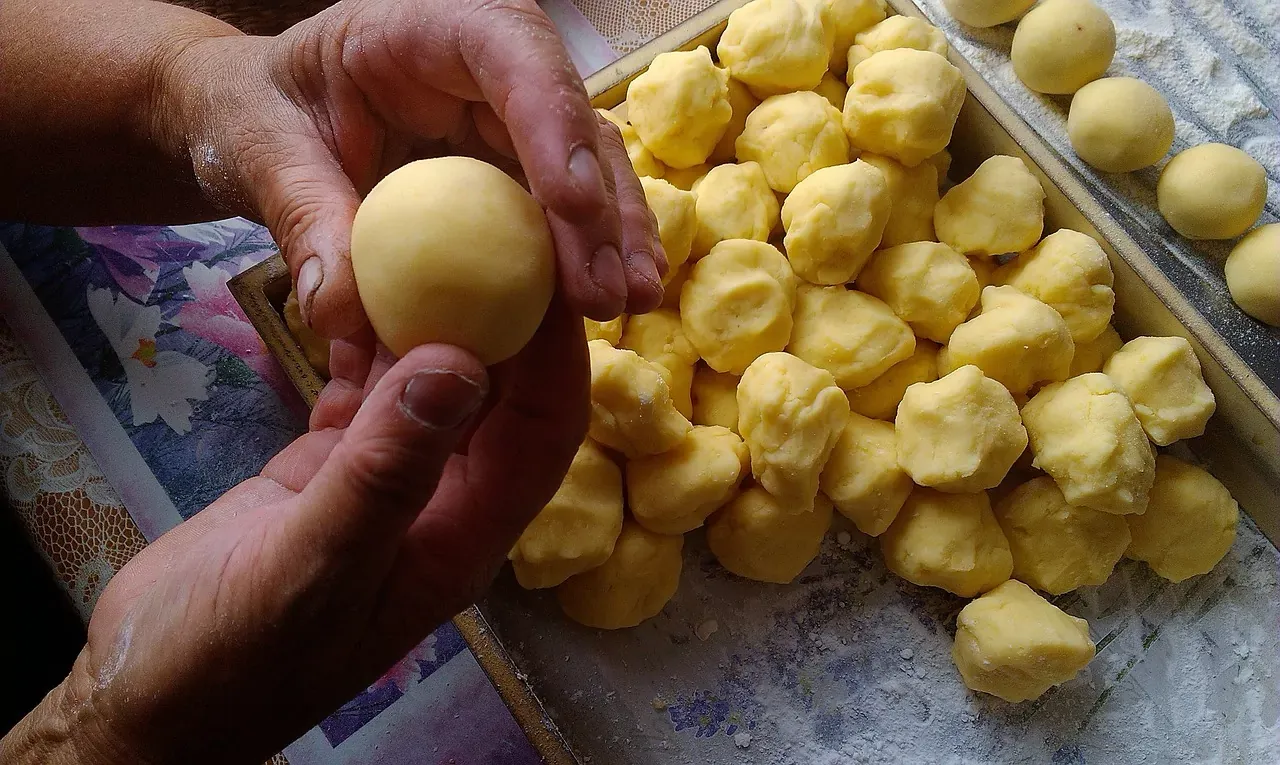 Hands holding a round, light yellow potato dumpling above a tray filled with many similar irregular dough pieces dusted with