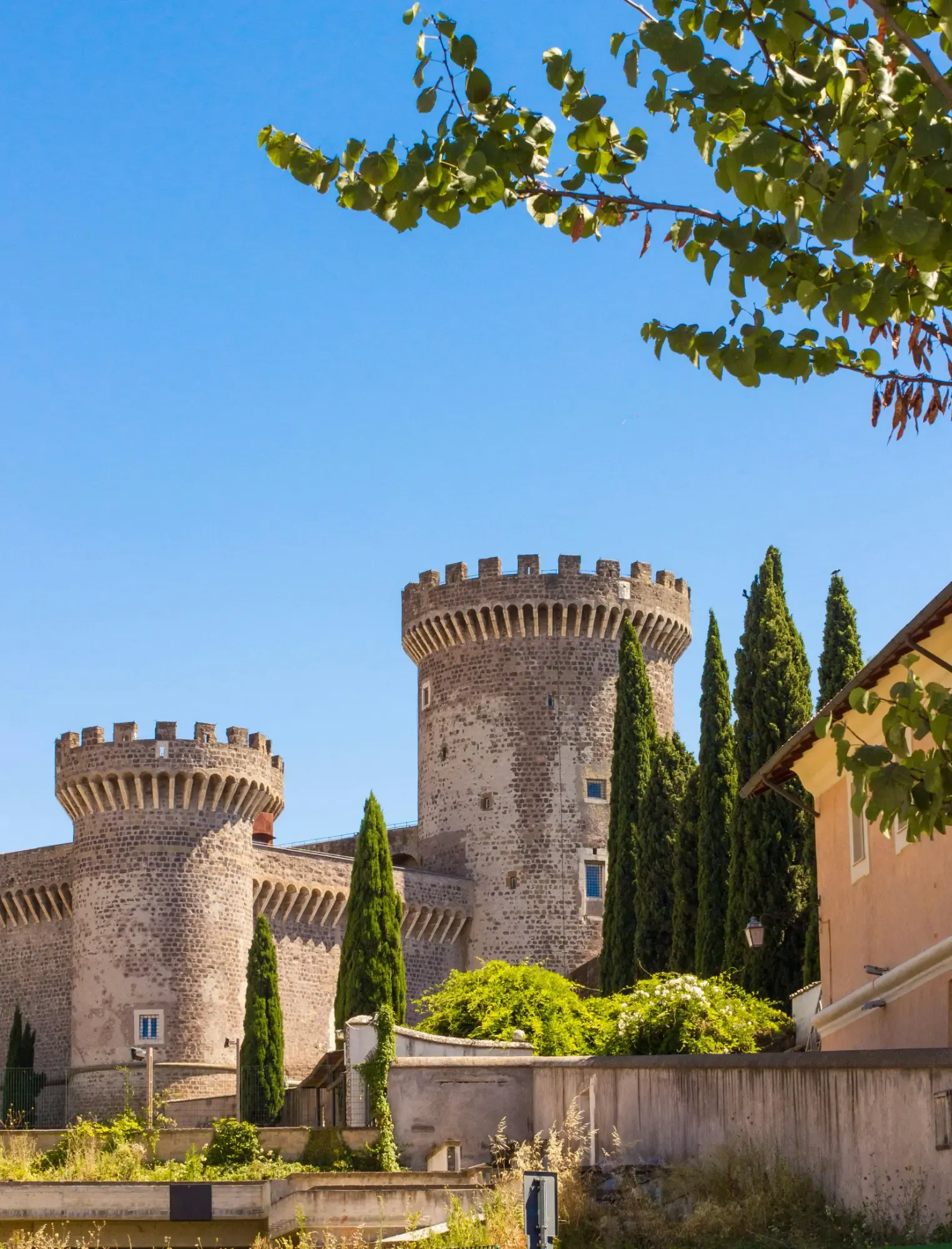 Two stone towers of Rocca Pia castle in Tivoli against a clear blue sky, with cypress trees nearby.