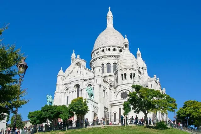 The white Sacré-Cœur Basilica in Paris stands prominently against a clear blue sky, with people on a grassy hill in front.