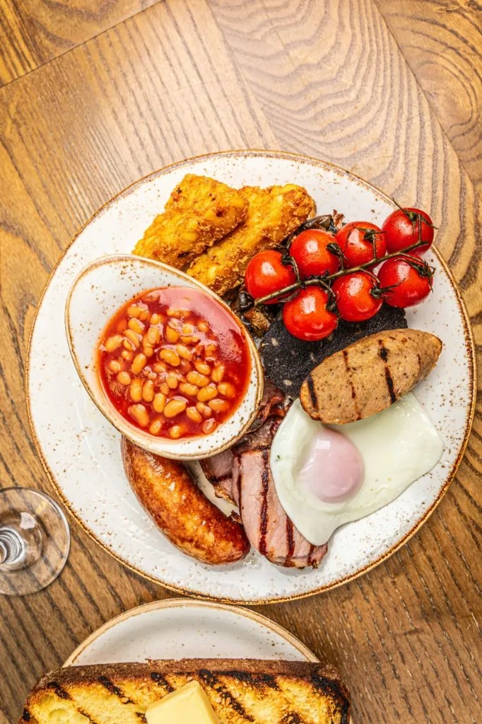 A full English breakfast on a white plate with baked beans, hash browns, cherry tomatoes, black pudding, sausage, bacon, and