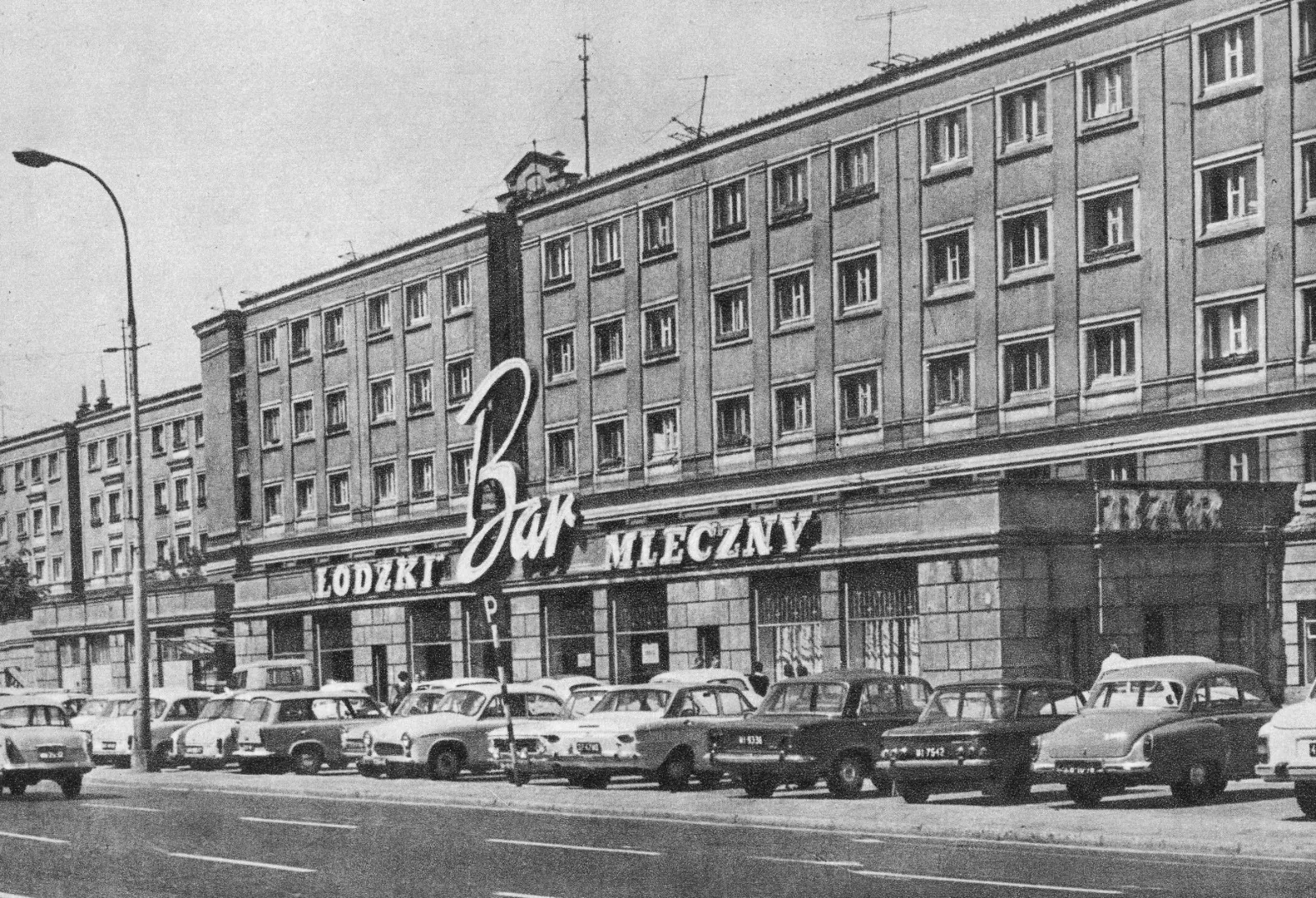 Street view of a Bar Mleczny building in Warsaw with its large sign. Several classic cars are parked along the curb.