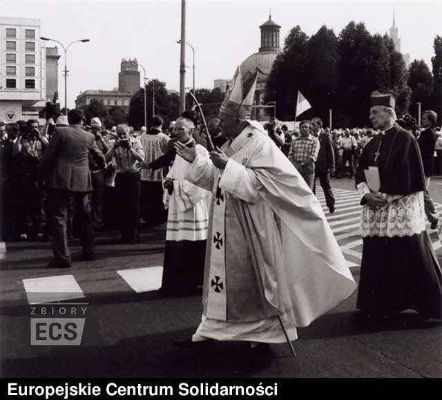 Pope John Paul II in full vestments walks across a street during a procession in Warsaw, surrounded by people.